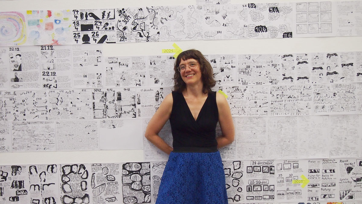 Simone lehnt an einer Wand, die voll beklebt ist mit vielen verschiedenen Zeichnungen in mehrern Reihen. Sie rägt ein schwarzes schulterfreies schwarzes Oberteil und einen blauen Rock. Sie trägt eine Brilel un hat mittellanges gewelltes Haar.