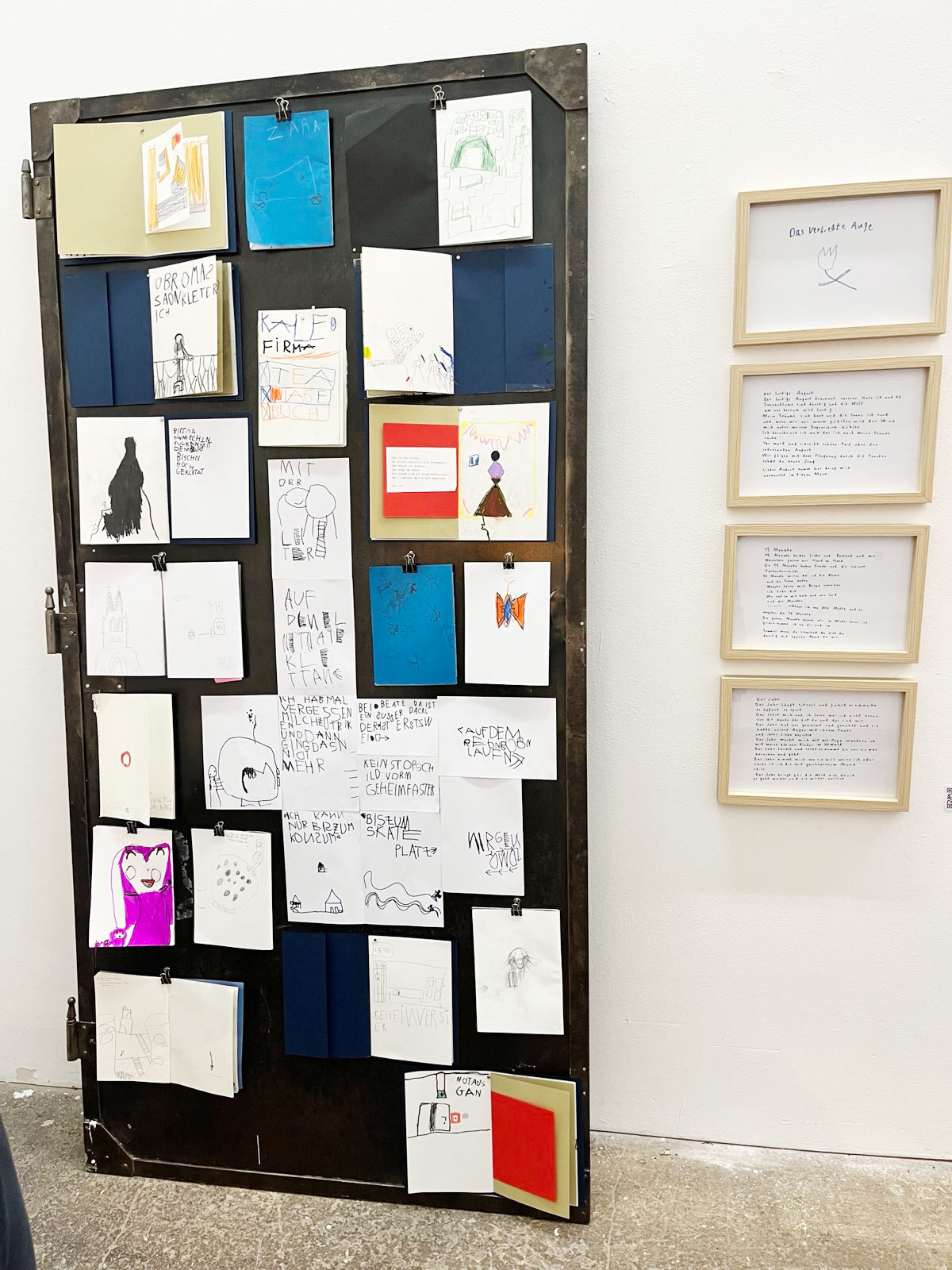 Eine dunkle Eisentühr lehnt an einer weißen Wand. An ihr sind viele kleine Hefte befestigt. man sieht Kinderzeichnungen und krakelige Schrift. Daneben 4 Rahmen übereinander mit handgeschriebenen Texten.