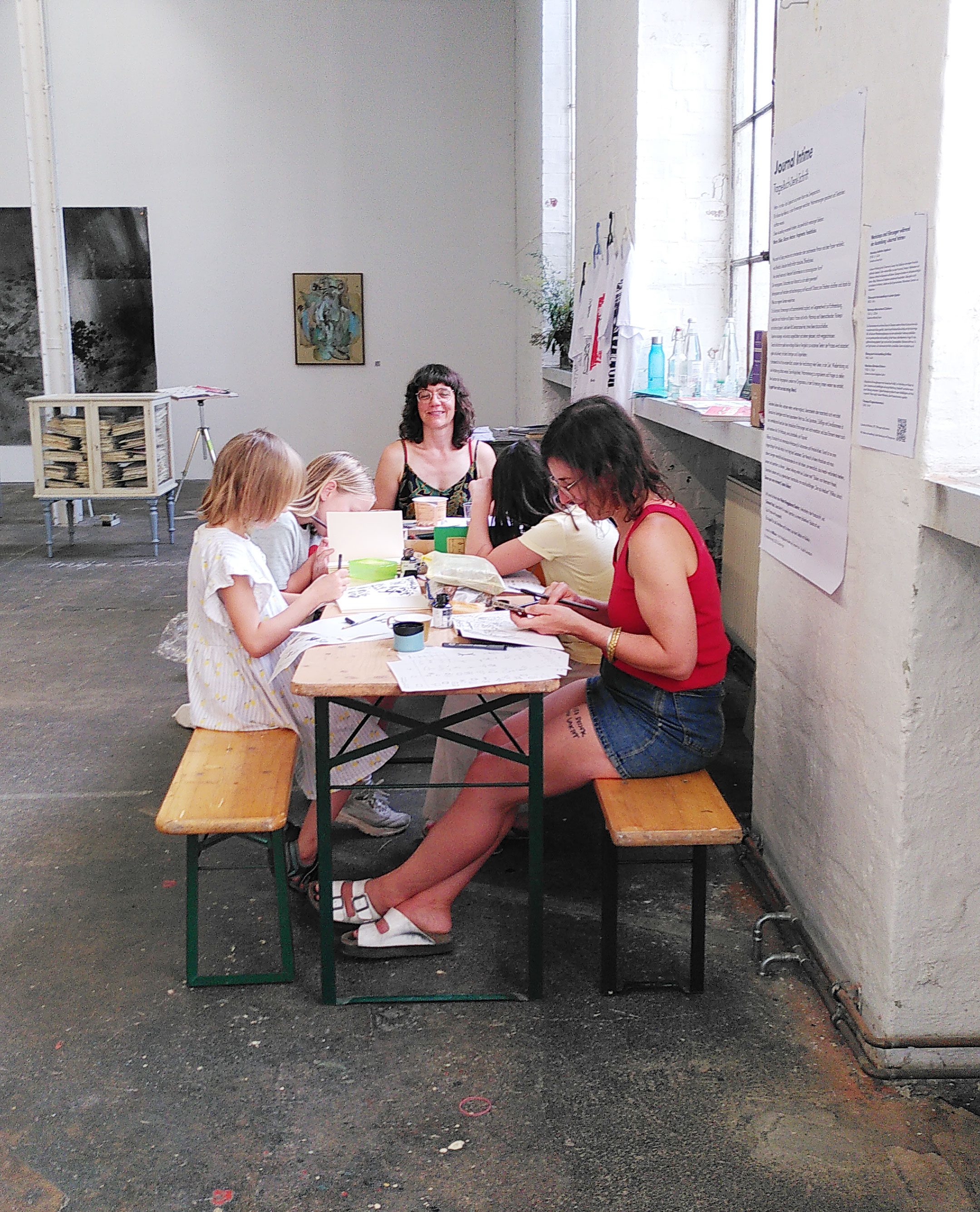 3 Kinder und 1 Frau sitzen mit Simone an einer Bierbank in einem hohen Ausstellungsraum. Sie zeichnen. Auf dem Tisch sind viele Papiere und Tusche. Simone sitzt am Kopf des Tisches und schaut auf die anderen. Im Hintergrund ein großer weißer Saal mit Bildern an der Wand.