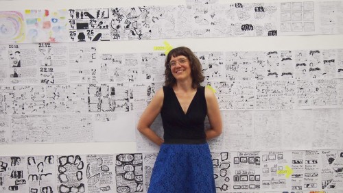 Simone lehnt an einer Wand, die voll beklebt ist mit vielen verschiedenen Zeichnungen in mehrern Reihen. Sie rägt ein schwarzes schulterfreies schwarzes Oberteil und einen blauen Rock. Sie trägt eine Brilel un hat mittellanges gewelltes Haar.