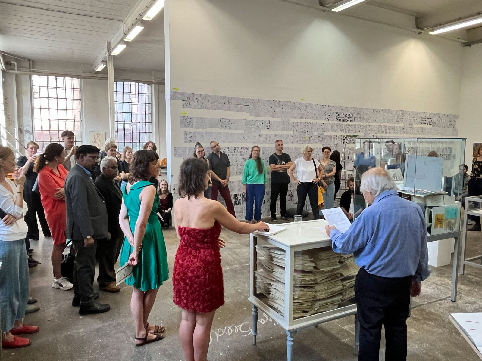 Foto eines sehr hohen Ausstellungsraums mit weißen Wänden und großen Industriefenstern. Im Vordergrund steht ein älterer Mann vor einer antiken weißen Vitrine. Er spricht gerade und hält ein Blatt dabei in der Hand. Daneben und dahinter stehen sehr viele Leute und hören ihm dabei zu. Im Hintergrund an einer großen Wand sind viele Zeichnungen an der Wand geklebt. In der Mitte des Raumes stehen Vitrinen mit vielen Büchern und Zeichnungen gefüllt.