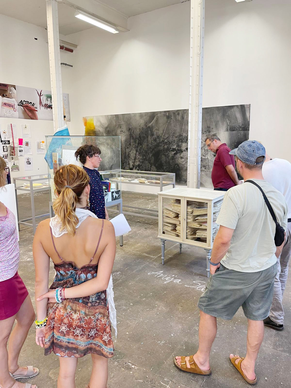 Eine kleine Gruppe Menschen steht im Ausstellungsraum vor einer antiken Vitrine. Simone steht am nahsten und schaut darauf. Im Hintergund zwei große weiße Säulen und ein großes graues Bild an der Wand.